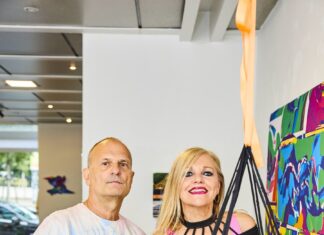 STATION ROSE: Delivering Digital Art since 1988 / Kulturforum London Das Bild zeigt die die Künstlerin Elisa Rose & Komponist Gary Danner in einer Porträtaufnahme. Sie blickt frontal in die Kamera. Sie stehen in einer Galerie mit einer Textilskulptur im Vordergrund und einem bunten Gemälde auf der rechten Seite. Elisa Rose trägt ein schwarzes Oberteil mit hellrosa BH und lächelt in die Kamera. Gary Danner trägt einen Batik-T-Shirt mit Cargo Hose. Er hat seine Hand in der Hosentasche und schaut intensiv in die Kamera.