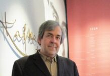 Kafkas letzte Reise / Kulturforum Prag Das Bild zeigt Manfred Müller in Porträtaufnahme – ein weißer Mann in Anzugsjacke mit grauen Haaren. Hinter ihm ist an der Wand der Schriftzug „Kafka“ sowie ein rotes Plakat mit musealer Zeitleiste, beginnend beim Jahr 1924 zu erkennen.