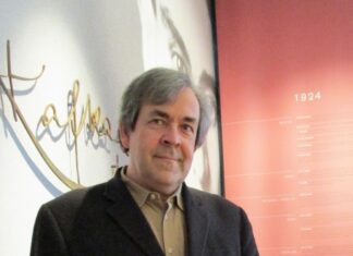 Kafkas letzte Reise / Kulturforum Prag Das Bild zeigt Manfred Müller in Porträtaufnahme – ein weißer Mann in Anzugsjacke mit grauen Haaren. Hinter ihm ist an der Wand der Schriftzug „Kafka“ sowie ein rotes Plakat mit musealer Zeitleiste, beginnend beim Jahr 1924 zu erkennen.