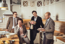 Konzert Candlelight Ficus/Botschaft Doha Das Bild zeigt die Band Candlelight Ficus: vier Männer mit grauen Anzügen stehen oder sitzen in einer weissen Küche und unterhalten sich. Das Outfit der Männer und die Küche wirken retro.