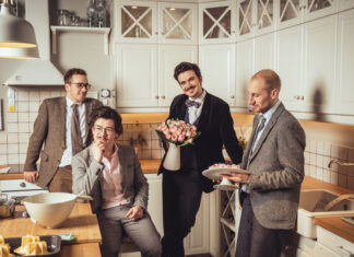 Konzert Candlelight Ficus/Botschaft Doha Das Bild zeigt die Band Candlelight Ficus: vier Männer mit grauen Anzügen stehen oder sitzen in einer weissen Küche und unterhalten sich. Das Outfit der Männer und die Küche wirken retro.