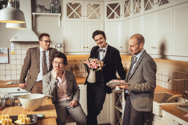 Das Bild zeigt die Band Candlelight Ficus: vier Männer mit grauen Anzügen stehen oder sitzen in einer weissen Küche und unterhalten sich. Das Outfit der Männer und die Küche wirken retro.