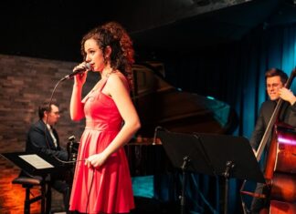 The Sound of Her / Kulturforum Berlin Das Bild zeigt eine Sängerin mit lockigem Haar. Sie trägt ein leuchtend rotes Kleid und hält ein Mikrofon in der Hand, während sie auf einer Bühne singt. Im Hintergrund sind ein Kontrabassist und ein Pianist zu sehen. Die Bühne ist in warmes Licht getaucht, und der Hintergrund besteht aus dunklen Vorhängen und einer Ziegelwand.