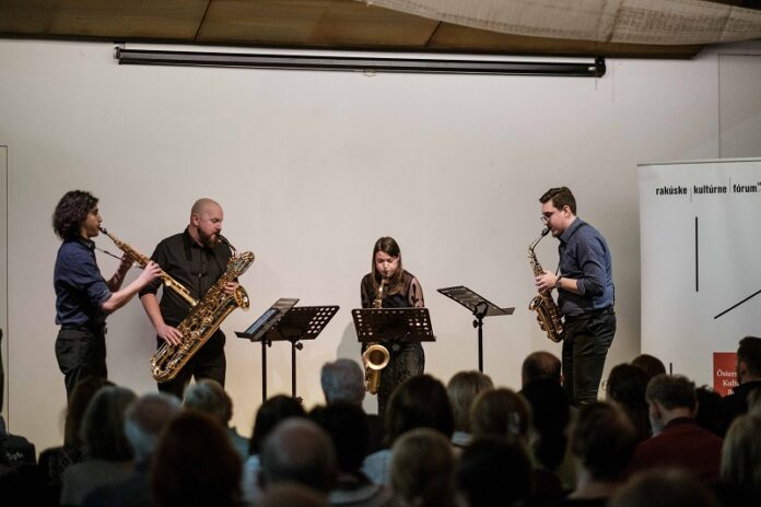 Das Bild zeigt das lächelnde „Spectrum Saxophonquartett“ vor einer weißen Wand. Alle sind dunkel angezogen und halten ihre Instrumente.