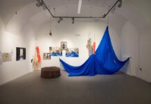 How are you in Celje? How are you in Graz?/ Kulturforum Ljubljana Das Bild zeigt einen weiß gestrichenen Ausstellungsraum, dessen Wände mit Fotografien und Kunstwerken dekoriert sind. Ein blaues drapiertes Tuch, das von der Decke hängt, nimmt den Großteil des Raums auf. Links daneben stehen bunte Flaggen und Stangen und auf dem Boden befindet sich ein gepolstertes rundes Möbelstück.