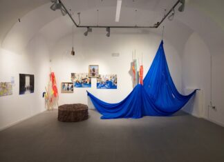 How are you in Celje? How are you in Graz?/ Kulturforum Ljubljana Das Bild zeigt einen weiß gestrichenen Ausstellungsraum, dessen Wände mit Fotografien und Kunstwerken dekoriert sind. Ein blaues drapiertes Tuch, das von der Decke hängt, nimmt den Großteil des Raums auf. Links daneben stehen bunte Flaggen und Stangen und auf dem Boden befindet sich ein gepolstertes rundes Möbelstück.