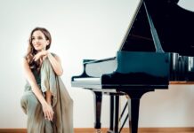 Klavierkonzert mit Mitra Kotte / Kulturforum London Das Bild zeigt Mitra Kotte, die links auf einem Klavierstuhl sitzt, mit einem Bein angewinkelt und lachend. Rechts ist das Klavier zu sehen.