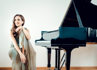 Klavierkonzert mit Mitra Kotte / Kulturforum London Das Bild zeigt Mitra Kotte, die links auf einem Klavierstuhl sitzt, mit einem Bein angewinkelt und lachend. Rechts ist das Klavier zu sehen.