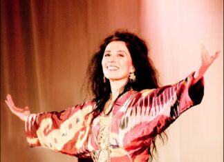 Abendkonzert / Kulturforum London Das Bild zeigt Sängerin Timna Brauer mit ausgebreiteten Armen. Sie trägt ein rotes, gemustertes Oberteil und hat lange, dunkle Haare. Ihr Gesichtsausdruck ist einladend und freundlich.