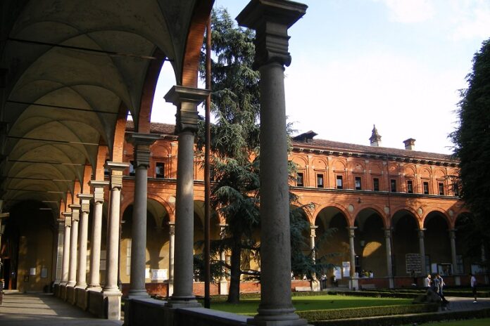 Das Bild zeigt den Hof von di Universität Milano Università degli Studi. Auf der linken Seite des Bilds sind Gänge mit hohen Bögen ersichtlich, während die rechte Seite mit dem Garten einen starken Kontrast bildet.