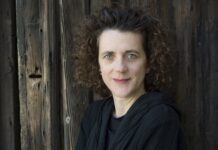 Radio France & Olga Neuwirth / Kulturforum Paris Das Bild zeigt eine in Schwarz gekleidete Olga Neuwirth, die vor einer Holzwand steht. Sie hat braune, kurze, lockige Haare und hellblaue Augen.