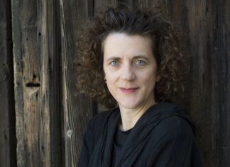 Radio France & Olga Neuwirth / Kulturforum Paris Das Bild zeigt eine in Schwarz gekleidete Olga Neuwirth, die vor einer Holzwand steht. Sie hat braune, kurze, lockige Haare und hellblaue Augen.