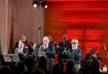 Nichts ist definitiv / Kulturforum Prag Das Bild zeigt vier Musiker mit Streichinstrumenten im hinteren und drei Schauspieler und Schauspielerinnen im vorderen Teil der Bühne. Vorne ist das Publikum in einem dunklen Saal zu sehen, die Bühne ist rot beleuchtet.