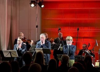 Nichts ist definitiv / Kulturforum Prag Das Bild zeigt vier Musiker mit Streichinstrumenten im hinteren und drei Schauspieler und Schauspielerinnen im vorderen Teil der Bühne. Vorne ist das Publikum in einem dunklen Saal zu sehen, die Bühne ist rot beleuchtet.