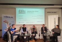 Smart Cities / Kulturforum Warschau Das Foto zeigt eine Podiumsdiskussion, bei der fünf Personen auf einer Bühne sitzen. Im Hintergrund ist eine Präsentationsfolie mit dem Titel „Smart Cities: Warsaw and Vienna in Dialogue“ zu sehen. Darauf stehen die Namen der Teilnehmerinnen und Teilnehmer sowie des Moderators. Links steht ein Banner mit dem Logo von „Smart City PW“, das bunte Symbole von Gebäuden, einem Flugzeug und anderen Elementen einer Stadt zeigt. Rechts ist ein Banner mit dem Logo des Österreichischen Kulturforums zu sehen. Die fünf Personen sitzen auf grauen Sesseln, vor ihnen stehen zwei niedrige Tische mit Glaskaraffen und Gläsern. Die Teilnehmerinnen und Teilnehmer sind formell gekleidet.