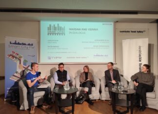Smart Cities / Kulturforum Warschau Das Foto zeigt eine Podiumsdiskussion, bei der fünf Personen auf einer Bühne sitzen. Im Hintergrund ist eine Präsentationsfolie mit dem Titel „Smart Cities: Warsaw and Vienna in Dialogue“ zu sehen. Darauf stehen die Namen der Teilnehmerinnen und Teilnehmer sowie des Moderators. Links steht ein Banner mit dem Logo von „Smart City PW“, das bunte Symbole von Gebäuden, einem Flugzeug und anderen Elementen einer Stadt zeigt. Rechts ist ein Banner mit dem Logo des Österreichischen Kulturforums zu sehen. Die fünf Personen sitzen auf grauen Sesseln, vor ihnen stehen zwei niedrige Tische mit Glaskaraffen und Gläsern. Die Teilnehmerinnen und Teilnehmer sind formell gekleidet.