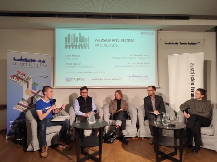 Das Foto zeigt eine Podiumsdiskussion, bei der fünf Personen auf einer Bühne sitzen. Im Hintergrund ist eine Präsentationsfolie mit dem Titel „Smart Cities: Warsaw and Vienna in Dialogue“ zu sehen. Darauf stehen die Namen der Teilnehmerinnen und Teilnehmer sowie des Moderators. Links steht ein Banner mit dem Logo von „Smart City PW“, das bunte Symbole von Gebäuden, einem Flugzeug und anderen Elementen einer Stadt zeigt. Rechts ist ein Banner mit dem Logo des Österreichischen Kulturforums zu sehen. Die fünf Personen sitzen auf grauen Sesseln, vor ihnen stehen zwei niedrige Tische mit Glaskaraffen und Gläsern. Die Teilnehmerinnen und Teilnehmer sind formell gekleidet.