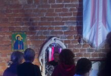 Ausstellung: Transposition: Kuratorische Narrative / Kulturforum Warschau Das Bild zeigt Besucher:innen in einem Ausstellungsraum. Der Raum ist schwach beleuchtet. Im Vordergrund sind Personen zu sehen, die mit dem Rücken zur Kamera stehen und die Ausstellung betrachten. Im Hintergrund ist eine Ziegelwand erkennbar. In der rechten oberen Ecke hängt eine künstlerische Stoffflagge in den Farben Blau, Rosa und Weiß. In der Mitte des Raumes befindet sich ein Spiegel, in dem sich das Gesicht einer Person spiegelt, die die Ausstellung betrachtet. Der Spiegel ist mit kleinen Fotografien beklebt. Auf der linken Seite an der Wand hängt ein Gemälde, das die Muttergottes mit dem Kind auf grünem Hintergrund darstellt. Die Farben des Gemäldes sind Blau, Gold und Orange.