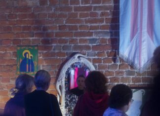 Ausstellung: Transposition: Kuratorische Narrative / Kulturforum Warschau Das Bild zeigt Besucher:innen in einem Ausstellungsraum. Der Raum ist schwach beleuchtet. Im Vordergrund sind Personen zu sehen, die mit dem Rücken zur Kamera stehen und die Ausstellung betrachten. Im Hintergrund ist eine Ziegelwand erkennbar. In der rechten oberen Ecke hängt eine künstlerische Stoffflagge in den Farben Blau, Rosa und Weiß. In der Mitte des Raumes befindet sich ein Spiegel, in dem sich das Gesicht einer Person spiegelt, die die Ausstellung betrachtet. Der Spiegel ist mit kleinen Fotografien beklebt. Auf der linken Seite an der Wand hängt ein Gemälde, das die Muttergottes mit dem Kind auf grünem Hintergrund darstellt. Die Farben des Gemäldes sind Blau, Gold und Orange.