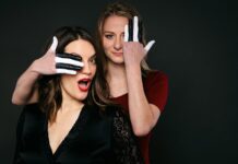 Dance, Dance, Otherwise We Are Lost / Kulturforen Washington und New York Das Bild zeigt die Sängerin Klaudia Tandl und die Pianistin Gisela Jöbstl. Tandl trägt ein schwarzes Kleid und einen knallroten Lippenstift, der gut zu ihren schulterlangen braunen Haaren passt. Hinter ihr steht die blonde Jöbstl, die ein rotes Shirt trägt und mit einer Hand ihr eigenes und mit der anderen das Auge der Sängerin bedeckt. Ihre Hände sind schwarz-weiß, wie Klaviertasten, bemalt.