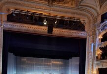 Johann Strauss Abend / Kulturforum Zagreb Das Bild zeigt Irene Suchy auf der Bühne während ihres Vortrags. Weil das Foto von der Ferne gemacht wurde, kann man sie nicht gut erkennen, aber sie trägt eine schwarze Hose und eine lange rote Jacke. Hinter ihr ist das Orchester in einem Halbkreis aufgestellt und wird mit kaltem blauen Licht beleuchtet. In der vorderen Hälfte des Bilds sind die Köpfe des sitzenden Publikums zu sehen. Der Konzertsaal ist weiß mit vielen goldenen Akzenten.