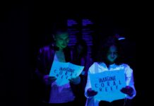 Imagine Dignity /Kulturforum Belgrad Das Bild zeigt zwei Frauen im Dunkeln, die auf ein fluoreszierendes Schild schauen, auf dem Coral Reef steht.