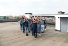 Rhythm is a Dancer / Kulturforum Berlin Das Bild zeigt eine Gruppe von Menschen, die auf einem Flachdach in einer synchronen Pose stehen. Sie sind in einer gestaffelten Formation angeordnet und heben beide Arme nach oben, die Handflächen zueinander gerichtet. Die Gruppe besteht aus Personen unterschiedlicher Geschlechter. Im Hintergrund sind Wohnhäuser mit roten Dächern sowie ein Industriegebäude mit Schornsteinen zu sehen. Der Himmel ist leicht bewölkt, und die Szene wirkt ruhig und konzentriert.