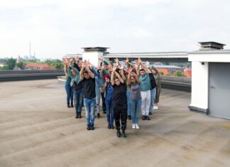 Rhythm is a Dancer / Kulturforum Berlin Das Bild zeigt eine Gruppe von Menschen, die auf einem Flachdach in einer synchronen Pose stehen. Sie sind in einer gestaffelten Formation angeordnet und heben beide Arme nach oben, die Handflächen zueinander gerichtet. Die Gruppe besteht aus Personen unterschiedlicher Geschlechter. Im Hintergrund sind Wohnhäuser mit roten Dächern sowie ein Industriegebäude mit Schornsteinen zu sehen. Der Himmel ist leicht bewölkt, und die Szene wirkt ruhig und konzentriert.