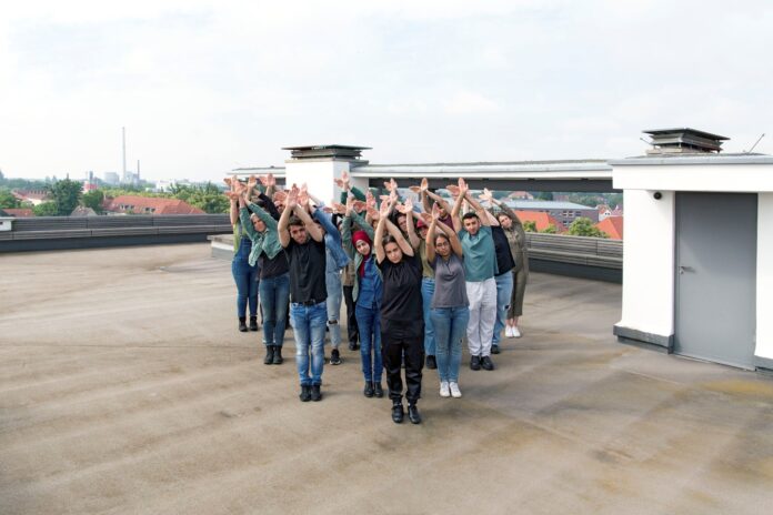 Das Bild zeigt eine Gruppe von Menschen, die auf einem Flachdach in einer synchronen Pose stehen. Sie sind in einer gestaffelten Formation angeordnet und heben beide Arme nach oben, die Handflächen zueinander gerichtet. Die Gruppe besteht aus Personen unterschiedlicher Geschlechter. Im Hintergrund sind Wohnhäuser mit roten Dächern sowie ein Industriegebäude mit Schornsteinen zu sehen. Der Himmel ist leicht bewölkt, und die Szene wirkt ruhig und konzentriert.