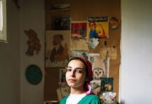 Finding Motherland – Franzi Kreis / Österreichisches Kulturforum Kairo Das Bild zeigt eine junge ägyptische Frau, die auf ihrem Bett kniet. Sie trägt eine blaue Jeans, eine grüne Jacke über einem weißen T-Shirt und ein rotes Kopftuch über ihre schwarzen Haare. Der Blick der jungen Frau ist direkt in die Kamera gerichtet. Ihre Hände stützt sie auf ihren Knien auf. Sie sitzt sehr aufrecht und zieht den Betrachter mit ihrem durchdringenden Blick in die Kulisse des Bildes. Im Hintergrund des Bildes befindet sich eine Wand, die gefüllt von Postern, Kunstwerken und Zetteln ist. Auch ein rosa Kuscheltier ist zu erkennen, was die Kindheit der jungen Frau thematisiert, eingebettet in den Raum, der vermutlich in dem Zimmer, was vermutlich einmal ihr eigenes Kinderzimmer war.