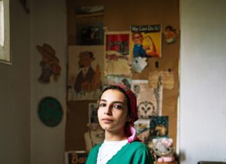 Finding Motherland – Franzi Kreis / Österreichisches Kulturforum Kairo Das Bild zeigt eine junge ägyptische Frau, die auf ihrem Bett kniet. Sie trägt eine blaue Jeans, eine grüne Jacke über einem weißen T-Shirt und ein rotes Kopftuch über ihre schwarzen Haare. Der Blick der jungen Frau ist direkt in die Kamera gerichtet. Ihre Hände stützt sie auf ihren Knien auf. Sie sitzt sehr aufrecht und zieht den Betrachter mit ihrem durchdringenden Blick in die Kulisse des Bildes. Im Hintergrund des Bildes befindet sich eine Wand, die gefüllt von Postern, Kunstwerken und Zetteln ist. Auch ein rosa Kuscheltier ist zu erkennen, was die Kindheit der jungen Frau thematisiert, eingebettet in den Raum, der vermutlich in dem Zimmer, was vermutlich einmal ihr eigenes Kinderzimmer war.