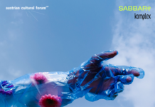 Ausstellungseröffnung Nile FLUX Das Bild zeigt eine in blaues Plastik eingewickelte Hand die vor dem Hintergrund des hellblauen Himmels in eine Richtung deutet. Auf dem Plastik befinden sich violette und lila farbene Blumen.