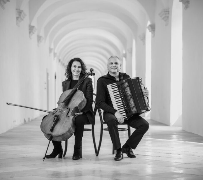 Das Bild zeigt das Duo sitzend, mit den jeweiligen Instrumenten in der Hand. Links sitzt die Cellistin Asja Valcic und hält ihr Cello, an ihrer rechten Seite ist der Akkordeonist Klaus Paier mit seinem Akkordeon abgebildet