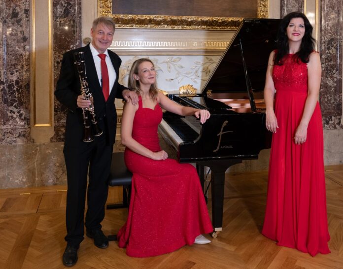 Das Bild zeigt das Trio vor einem Klavier. Die Pianistin und die Sopranistin in rotem Kleid. Der Musiker in schwarzem Anzug.