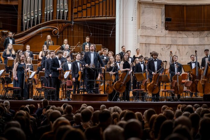 Das Bild zeigt eine große Orchesteraufführung in einem Konzertsaal. Die Musikerinnen und Musiker stehen auf der Bühne und verbeugen sich vor dem Publikum. Es ist ein jugendorientiertes Orchester zu sehen, mit vielen jungen Musikern in schwarzer Formalkleidung, darunter schwarze Anzüge und Kleider. Der Dirigent steht auf einem erhöhten Podium und hat eine Hand erhoben, um das Publikum zu begrüßen. Im Hintergrund sieht man einen Teil der Orgel des Saals, was auf eine große, prunkvolle Konzertlocation hinweist. Das Publikum ist im unteren Bildbereich sichtbar, einige Hände klatschen, was auf den Applaus und die Anerkennung für die Darbietung hinweist. Die Atmosphäre wirkt festlich und feierlich, mit einem Gefühl von Stolz und Erfolg nach der Aufführung.