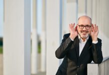 Pianistic Impressions from Vienna / Kulturforum Washington Das Bild zeigt den Pianisten Gottlieb Wallisch. Er lehnt an einer Wand, lacht und hat die Hände hinter den Ohren. Er trägt einen schwarzen Anzug und hat eine schwarze Brille auf. Er hat wenig Haare und lacht sehr herzlich.