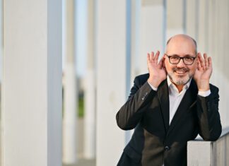 Pianistic Impressions from Vienna / Kulturforum Washington Das Bild zeigt den Pianisten Gottlieb Wallisch. Er lehnt an einer Wand, lacht und hat die Hände hinter den Ohren. Er trägt einen schwarzen Anzug und hat eine schwarze Brille auf. Er hat wenig Haare und lacht sehr herzlich.