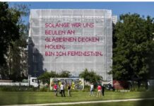 Solange – The Project / Botschaft Oslo Das Bild zeigt eines der Netze des Solange Projektes. Im Vordergrund ist eine grüne Wiese zu sehen auf der 6 Menschen in zwei Dreiergruppen zusammenstehen. Sie haben den Betrachtern den Rücken zugewandt und schauen in Richtung des Kunstwerkes. Mittig im Bild ist ein Baustellengerüst, eingerahmt von Bäumen, an dem das weiße Netz-Kunstwerk aufgespannt ist. In pinkem Kreuzstich ist in Großbuchstaben auf dem Netz der Spruch „Solange wir uns Beulen an gläsernen Decken holen, bin ich FeministIn“ aufgestickt.