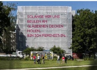 Solange – The Project / Botschaft Oslo Das Bild zeigt eines der Netze des Solange Projektes. Im Vordergrund ist eine grüne Wiese zu sehen auf der 6 Menschen in zwei Dreiergruppen zusammenstehen. Sie haben den Betrachtern den Rücken zugewandt und schauen in Richtung des Kunstwerkes. Mittig im Bild ist ein Baustellengerüst, eingerahmt von Bäumen, an dem das weiße Netz-Kunstwerk aufgespannt ist. In pinkem Kreuzstich ist in Großbuchstaben auf dem Netz der Spruch „Solange wir uns Beulen an gläsernen Decken holen, bin ich FeministIn“ aufgestickt.