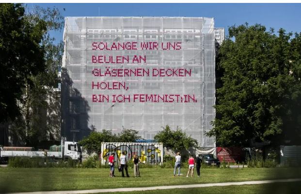 Das Bild zeigt eines der Netze des Solange Projektes. Im Vordergrund ist eine grüne Wiese zu sehen auf der 6 Menschen in zwei Dreiergruppen zusammenstehen. Sie haben den Betrachtern den Rücken zugewandt und schauen in Richtung des Kunstwerkes. Mittig im Bild ist ein Baustellengerüst, eingerahmt von Bäumen, an dem das weiße Netz-Kunstwerk aufgespannt ist. In pinkem Kreuzstich ist in Großbuchstaben auf dem Netz der Spruch „Solange wir uns Beulen an gläsernen Decken holen, bin ich FeministIn“ aufgestickt.