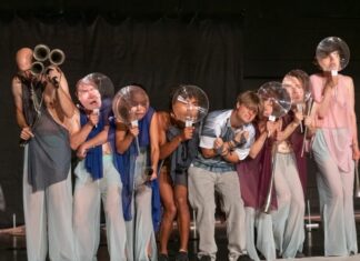 Eva Reiter – The Rise / Kulturforum Brüssel Das Bild zeigt eine Szene aus dem Theaterstück „The Rise“. Sieben Personen sind auf einem Podium zu sehen. Alle halten Glasstücke, die wie Seifenblasen aussehen, vor ihren Augen.