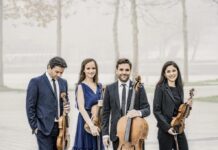 Minetti Quartett: Schubertiade / Kulturforum New York Das Bild zeigt das Minetti Quartett, bestehend aus vier Musikerinnen und Musikern, die mit ihren Streichinstrumenten im Freien posieren. Sie tragen elegante, formelle Kleidung – die beiden Frauen in einem blauen Kleid bzw. einem schwarzen Anzug, die beiden Männer in dunklen Anzügen mit Krawatten. Alle halten ihre Instrumente: zwei Violinen, eine Viola und ein Cello. Der Hintergrund ist neblig, mit kahlen Bäumen und einer verschwommenen Landschaft, was eine ruhige, fast verträumte Atmosphäre schafft. Die Gruppe steht entspannt beisammen, einige Mitglieder lächeln in die Kamera, während andere nach unten blicken.
