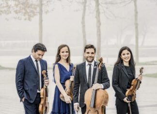 Minetti Quartett: Schubertiade / Kulturforum New York Das Bild zeigt das Minetti Quartett, bestehend aus vier Musikerinnen und Musikern, die mit ihren Streichinstrumenten im Freien posieren. Sie tragen elegante, formelle Kleidung – die beiden Frauen in einem blauen Kleid bzw. einem schwarzen Anzug, die beiden Männer in dunklen Anzügen mit Krawatten. Alle halten ihre Instrumente: zwei Violinen, eine Viola und ein Cello. Der Hintergrund ist neblig, mit kahlen Bäumen und einer verschwommenen Landschaft, was eine ruhige, fast verträumte Atmosphäre schafft. Die Gruppe steht entspannt beisammen, einige Mitglieder lächeln in die Kamera, während andere nach unten blicken.