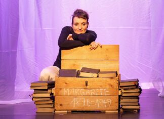 Leb‘ wohl, Bohème / Botschaft Pristina Das Bild zeigt die Schauspielerin Alice Schneider auf der Bühne hinter eine Kiste voller Bücher kniend.