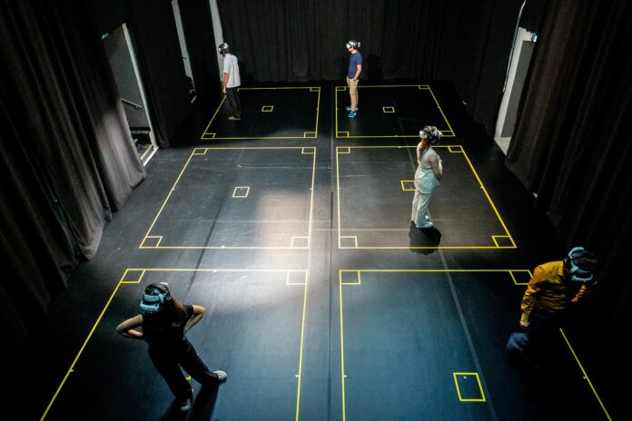 Das Bild zeigt sechs Personen in einem dunklen Raum mit VR-Headsets. Sie stehen jeweils in abgegrenzten Feldern, markiert durch gelbe Linien auf dem Boden. Der Raum ist von schwarzen Vorhängen umgeben, die Atmosphäre wirkt ruhig und konzentriert.