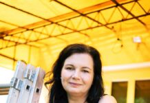 Generation Beta / Kulturforum Budapest Das Bild zeigt eine Frau vor ihrem Haus. Sie trägt ein schwarzes ärmelloses Kleid mit weissen Punkten und hat die Hände über einer Brüstung verschränkt. Sie hat dunkle lange Haare und lächelt in die Kamera.