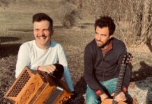 Konzert mit Jakob Steinkellner / Kulturforum London Das Bild zeigt Jakob Steinkellner auf der linken Seite, der eine Steirische Harmonika hält, während Severin Trogbacher auf der rechten Seite mit einer Gitarre zu sehen ist. Beide sitzen in der Sonne in einer natürlichen Umgebung, lachen und halten ihre Instrumente.