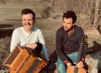 Konzert mit Jakob Steinkellner / Kulturforum London Das Bild zeigt Jakob Steinkellner auf der linken Seite, der eine Steirische Harmonika hält, während Severin Trogbacher auf der rechten Seite mit einer Gitarre zu sehen ist. Beide sitzen in der Sonne in einer natürlichen Umgebung, lachen und halten ihre Instrumente.