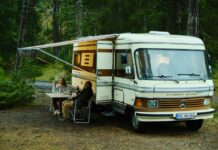 BFFB38 – Bolzano Film Festival Bozen / Kulturforum Mailand Das Bild zeigt eine Frau und einen Mann, die in der Natur vor einem altmodischen Wohnmobil sitzen. Es ist eine Szene aus dem Film Marianengraben.