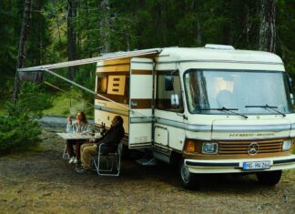 BFFB38 – Bolzano Film Festival Bozen / Kulturforum Mailand Das Bild zeigt eine Frau und einen Mann, die in der Natur vor einem altmodischen Wohnmobil sitzen. Es ist eine Szene aus dem Film Marianengraben.